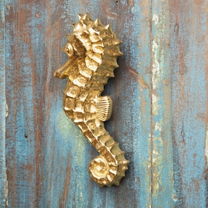 May include: A gold-coloured seahorse door knocker with a detailed design. The knocker is mounted on a blue wooden door.