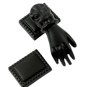 May include: Black metal door knocker shaped like a hand with a ring on the finger. The knocker is attached to a square base with a decorative border.