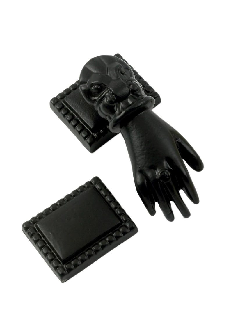 May include: Black metal door knocker shaped like a hand with a ring on the finger. The knocker is attached to a square base with a decorative border.