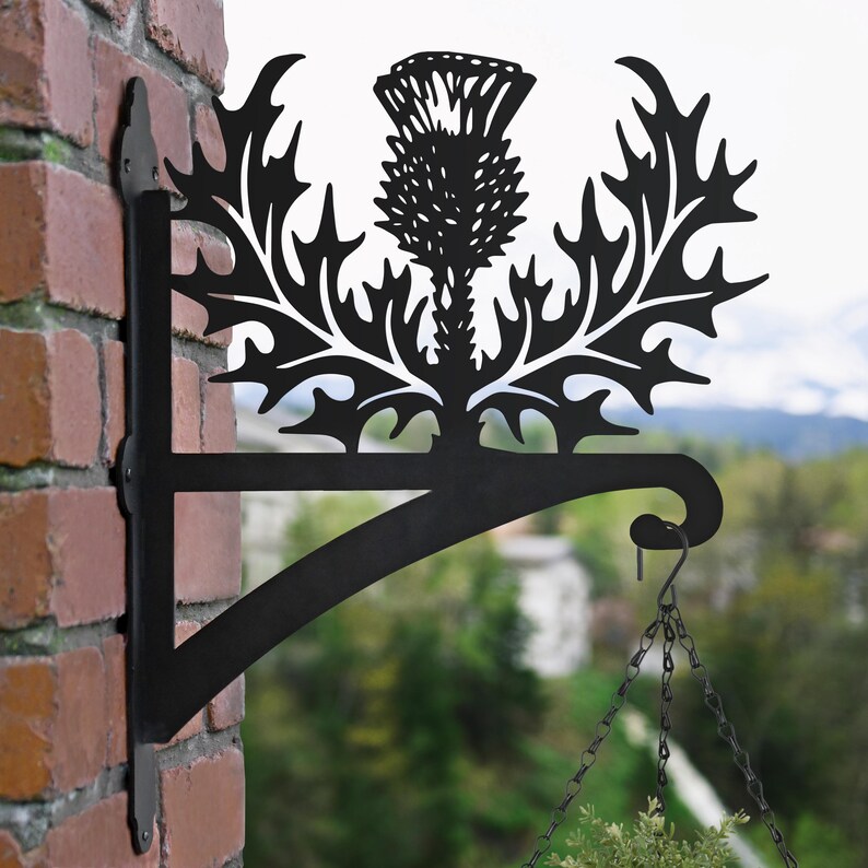 May include: Black metal plant hanger with a thistle design. The hanger is attached to a brick wall and has a chain hanging from the bottom.