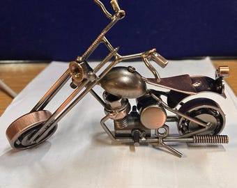 May include: A miniature motorcycle made of metal, with a silver and copper finish. The motorcycle has a single seat and a long, straight handlebar.