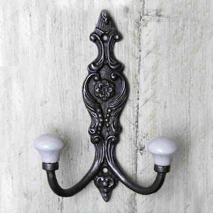 May include: A black cast iron double hook with ornate detailing and two white ceramic knobs. The hook is mounted on a white wooden wall.