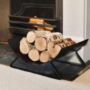 May include: A matte black metal log holder with a curved, open-trough design and angled support legs, filled with several pieces of light-coloured, textured birch firewood with visible bark and circular grain patterns on the ends.