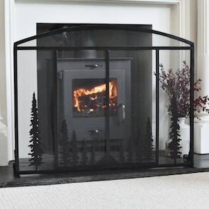 May include: A black metal fireplace screen featuring an arched top and a fine mesh panel. The lower section of the screen is decorated with a silhouette design of various pine trees across the front, providing a decorative barrier for the hearth.