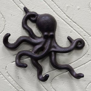 May include: A dark bronze octopus-shaped wall hook. The octopus has eight curling tentacles and a rounded head. The hook is mounted on a textured, off-white surface. The design is a nautical or coastal theme.