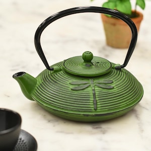 May include: Green cast iron teapot with a dragonfly design and a black handle. The teapot has a lid with a small knob on top.