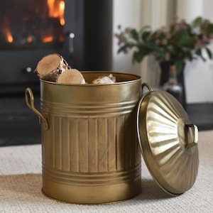 May include: A brass-toned metal log bin with a lid and handles, featuring vertical ribbed detailing. The lid is open, showing the inside filled with cut logs. The bin is on a neutral rug, with a fireplace visible in the background.