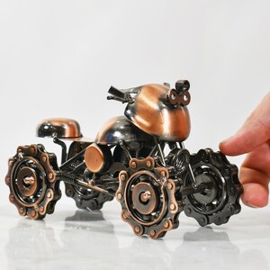 Puede incluir: Una peque&ntilde;a escultura de metal de color cobre de una motocicleta con ruedas de cadena. La motocicleta tiene una cabeza redondeada, similar a un casco, y un solo asiento.