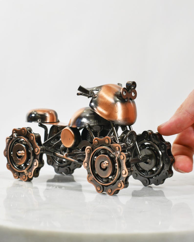 Puede incluir: Una peque&ntilde;a escultura de metal de color cobre de una motocicleta con ruedas de cadena. La motocicleta tiene una cabeza redondeada, similar a un casco, y un solo asiento.