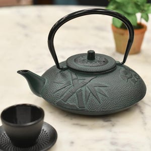 May include: A green cast iron teapot with a black handle and a bamboo design. The teapot is sitting on a white marble surface.