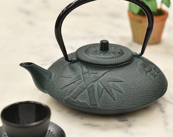May include: A green cast iron teapot with a black handle and a bamboo design. The teapot is sitting on a white marble surface.