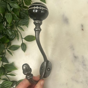May include: A decorative wall hook with a black and white striped ceramic knob. The hook is made of dark grey metal and features an acorn-shaped end. The hook is mounted on a white marble surface.