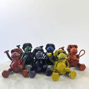 May include: A group of metal monkey sculptures in various colours. Each monkey is made from nuts and bolts, with shades of red, green, purple, blue, and yellow. The sculptures are displayed on a white surface.