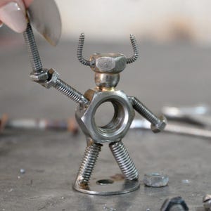 May include: A handcrafted metal sculpture of a figure holding an axe. The figure is constructed from nuts, bolts, and springs, with a circular body and a head made from a hex nut. The axe is held in the figure's raised arm.