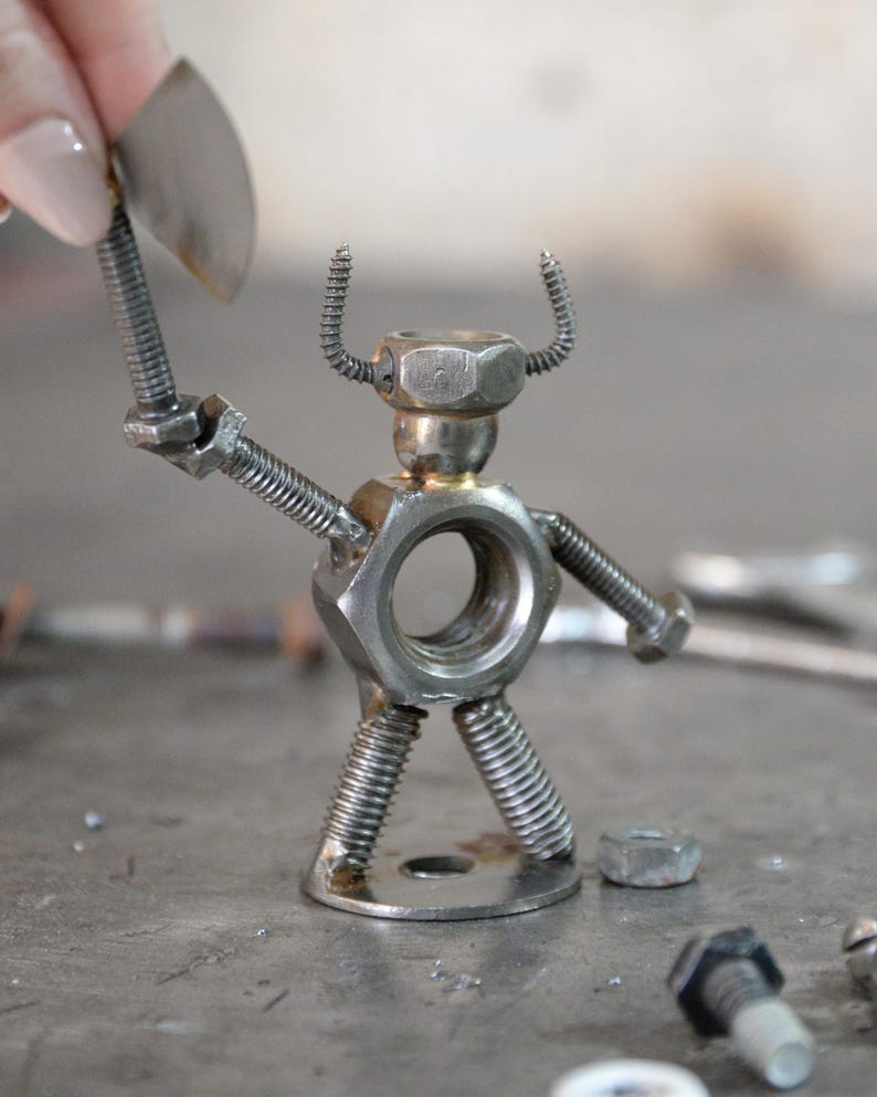 May include: A handcrafted metal sculpture of a figure holding an axe. The figure is constructed from nuts, bolts, and springs, with a circular body and a head made from a hex nut. The axe is held in the figure's raised arm.