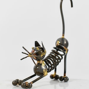 May include: A dark metal sculpture of a cat in a stretching pose. The body is formed by a coiled spring, with spherical metal beads for the head, paws, and tail base. The sculpture features thin wire whiskers and a long, upright curved tail.
