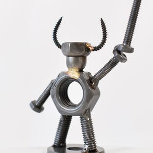 May include: A metal sculpture of a robot made from nuts, bolts, and screws. The robot has a large hexagonal nut for a torso, a smaller nut for a head, and curved screw horns. It holds a bolt in one hand, and has screw legs.