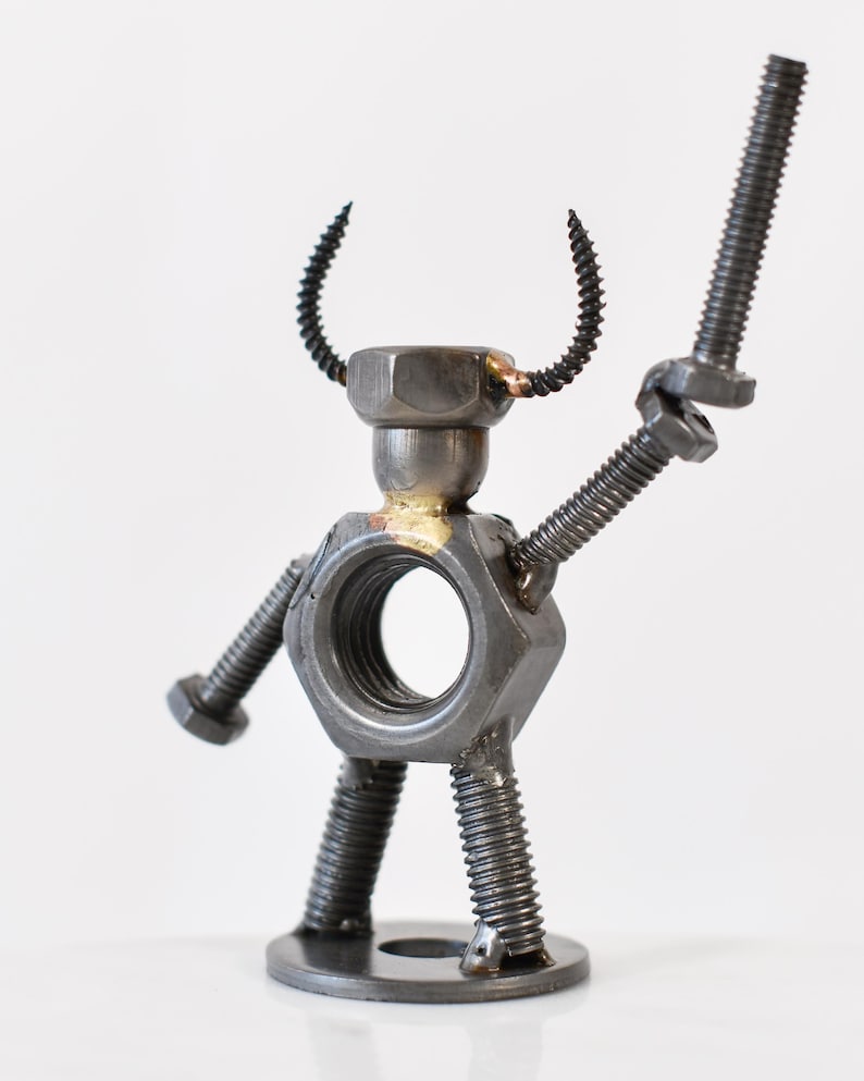 May include: A metal sculpture of a robot made from nuts, bolts, and screws. The robot has a large hexagonal nut for a torso, a smaller nut for a head, and curved screw horns. It holds a bolt in one hand, and has screw legs.