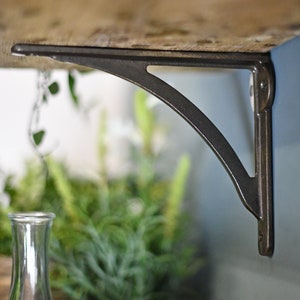 May include: A black metal shelf bracket with a curved design. The bracket is attached to a wooden shelf and is mounted to a wall.