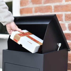 May include: A black, contemporary mailbox with a pitched roof, open to receive a white cardboard box. The box is sealed with brown tape and labelled "FRAGILE" in red. The mailbox is mounted on a brick wall.