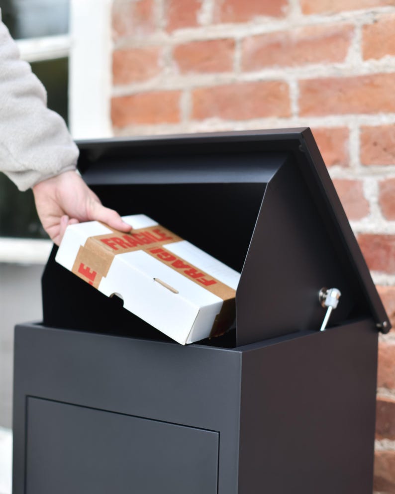 May include: A black, contemporary mailbox with a pitched roof, open to receive a white cardboard box. The box is sealed with brown tape and labelled "FRAGILE" in red. The mailbox is mounted on a brick wall.