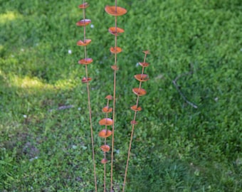 May include: Outdoor metal garden art featuring three vertical sculptures. Each sculpture has a series of circular, rust-colored discs attached to thin metal rods. The sculptures are set against a backdrop of green grass.