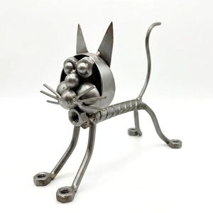 May include: A metal cat sculpture, handcrafted from nuts, bolts, and metal rods. The cat features a head with pointed ears and whiskers, a body, legs, and a curved tail. The sculpture has a silver finish. The cat's dimensions are approximately 20 cm in length.