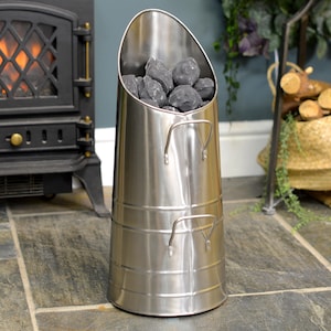 May include: A silver metal coal scuttle with two handles. The scuttle is filled with black coal briquettes. The scuttle is sitting on a tiled floor in front of a black fireplace.