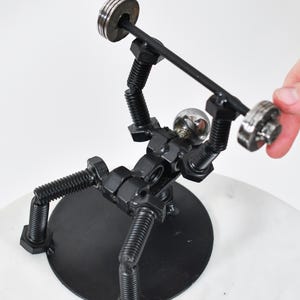May include: A black metal sculpture of a person lifting weights. The sculpture is made of nuts, bolts, and screws. The person is lifting a barbell with two weights on each end. The sculpture is standing on a black circular base.