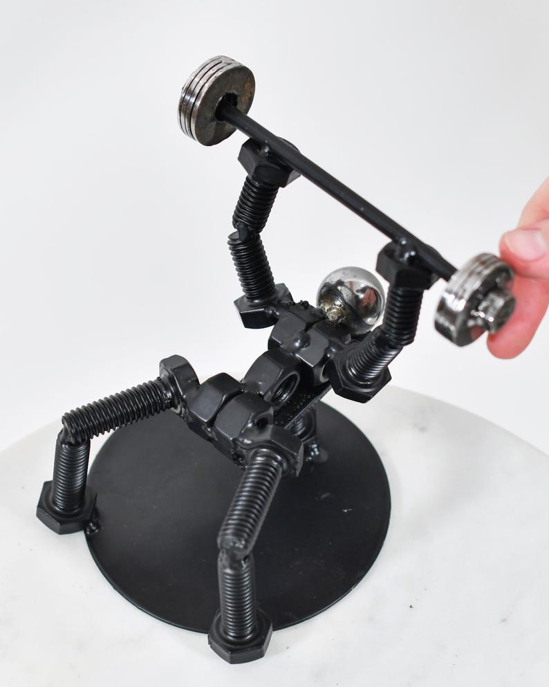 May include: A black metal sculpture of a person lifting weights. The sculpture is made of nuts, bolts, and screws. The person is lifting a barbell with two weights on each end. The sculpture is standing on a black circular base.