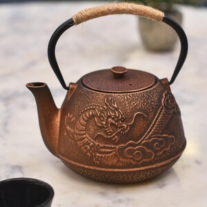 May include: A copper-coloured cast iron teapot with a dragon design and a rope handle. The teapot is sitting on a white marble surface.