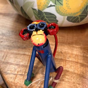 May include: A monkey figurine constructed from metal hardware components including blue threaded rods for limbs, red nuts for ears, and a blue cylindrical body. The face is painted yellow with red accents and black details, featuring dark blue nut eyes.