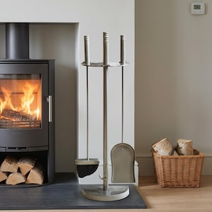 May include: A brushed silver metal fireplace tool set featuring a central stand with a circular base, holding a shovel, a poker, and a brush with black bristles. The tools have matching cylindrical handles and are arranged in a vertical rack.