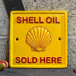 May include: A yellow metal sign with red lettering that reads "SHELL OIL SOLD HERE". The sign features a red and yellow stylised seashell logo.