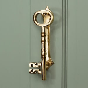 May include: A decorative brass key door knocker. The key-shaped knocker has an oval handle, a detailed stem, and a key bit. It is mounted on a light green door. The overall design is ornate and eye-catching.