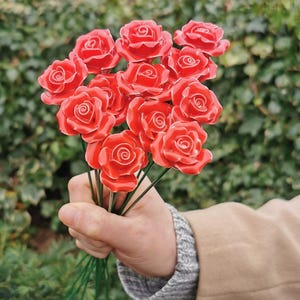 May include: A hand holding a bunch of red ceramic roses with green wire stems. The roses are glossy and detailed, set against a blurred green backdrop. The flowers have a realistic appearance.