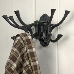 May include: Black cast iron coat rack with multiple curved hooks, mounted on a white wall. A brown and tan plaid shirt hangs from one of the hooks. The rack has a decorative, antler-like design.