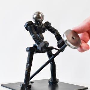 May include: A metal sculpture of a person lifting a weight. The sculpture is made of nuts, bolts, and other metal pieces. The person is standing on a black metal base.