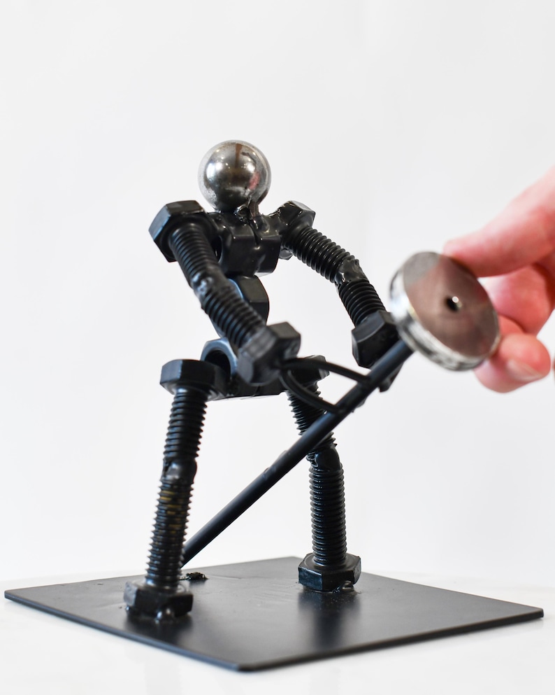 May include: A metal sculpture of a person lifting a weight. The sculpture is made of nuts, bolts, and other metal pieces. The person is standing on a black metal base.