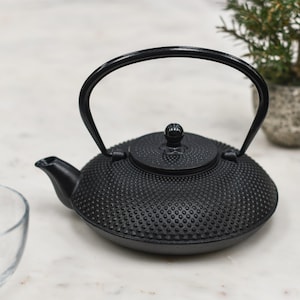 May include: Black cast iron teapot with a textured surface and a curved handle. The teapot has a small spout and a lid with a knob. The teapot is designed for brewing tea and has a classic, traditional appearance.