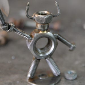May include: A handcrafted metal sculpture of a figure made from nuts, bolts, and screws. The figure has a hexagonal nut for a head, screw horns, and bolt arms. The legs are made from screws, and it stands on a metal base.