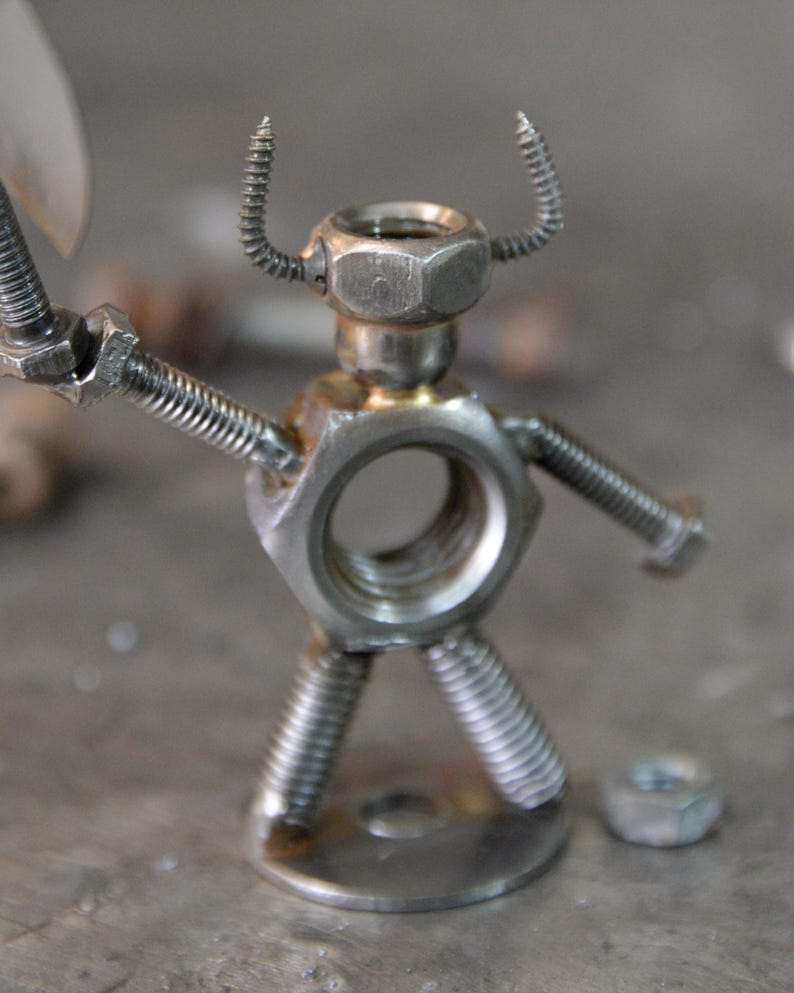 May include: A handcrafted metal sculpture of a figure made from nuts, bolts, and screws. The figure has a hexagonal nut for a head, screw horns, and bolt arms. The legs are made from screws, and it stands on a metal base.