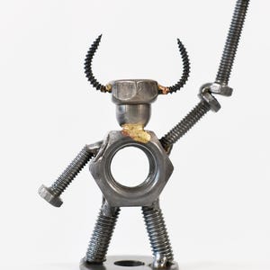 May include: A whimsical metal sculpture of a figure assembled from nuts, bolts, and screws. The figure has a nut for a torso, a bolt for an arm, and screw-shaped horns. The sculpture is on a white background.