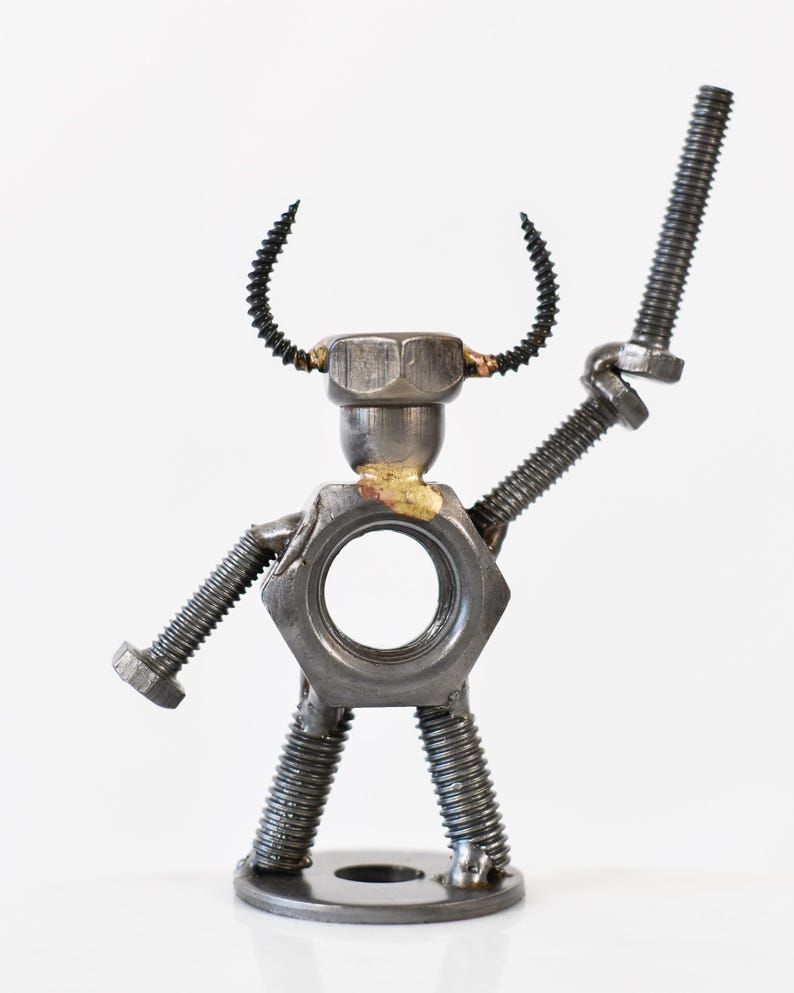 May include: A whimsical metal sculpture of a figure assembled from nuts, bolts, and screws. The figure has a nut for a torso, a bolt for an arm, and screw-shaped horns. The sculpture is on a white background.