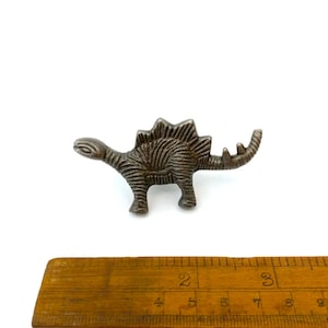 May include: A small, metallic Stegosaurus figurine with detailed ridged texture. The dinosaur is a muted silver colour and stands on four legs. A wooden ruler is in the foreground.