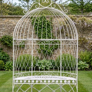 May include: An ivory-coloured, ornate metal garden bench with a domed top. The bench features intricate scrollwork and a diamond lattice design. It is set on a green lawn with a stone wall and greenery in the background.