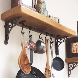 May include: A wooden shelf with black metal brackets and hooks. The shelf is mounted on a white wall and has various kitchen utensils hanging from the hooks, including a wooden spoon, a metal pot, and a frying pan. A small cat figurine is hanging from one of the hooks.