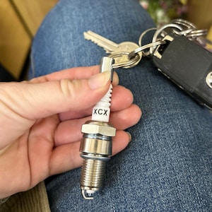 May include: A white spark plug keychain with the text "XCX" printed on it. The spark plug is attached to a silver key ring.
