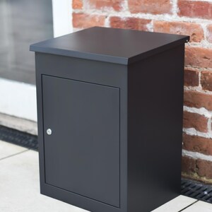May include: A dark grey, rectangular mailbox with a hinged door and a small silver handle. The mailbox is set against a red brick wall and a white tiled surface. The top of the mailbox is flat.