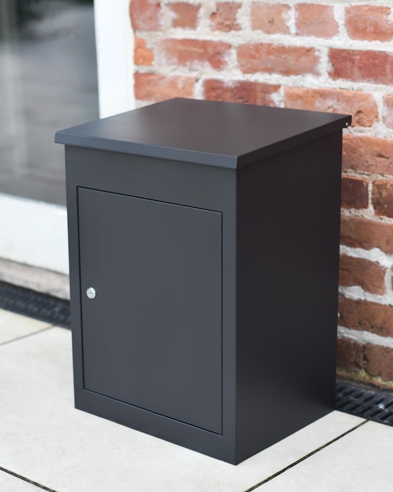 May include: A dark grey, rectangular mailbox with a hinged door and a small silver handle. The mailbox is set against a red brick wall and a white tiled surface. The top of the mailbox is flat.